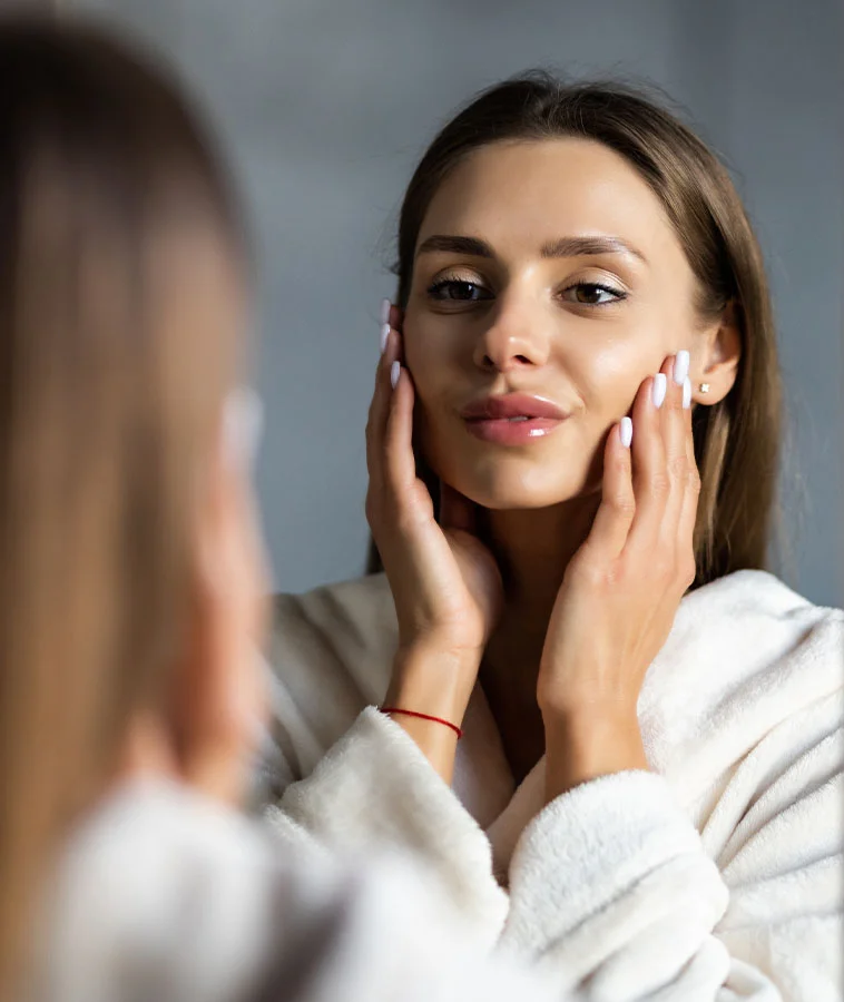 A young woman with long brown hair, wearing a white robe, touching her face and admiring her reflection in the mirror - Buccal Fat Removal Procedure in New York, NY