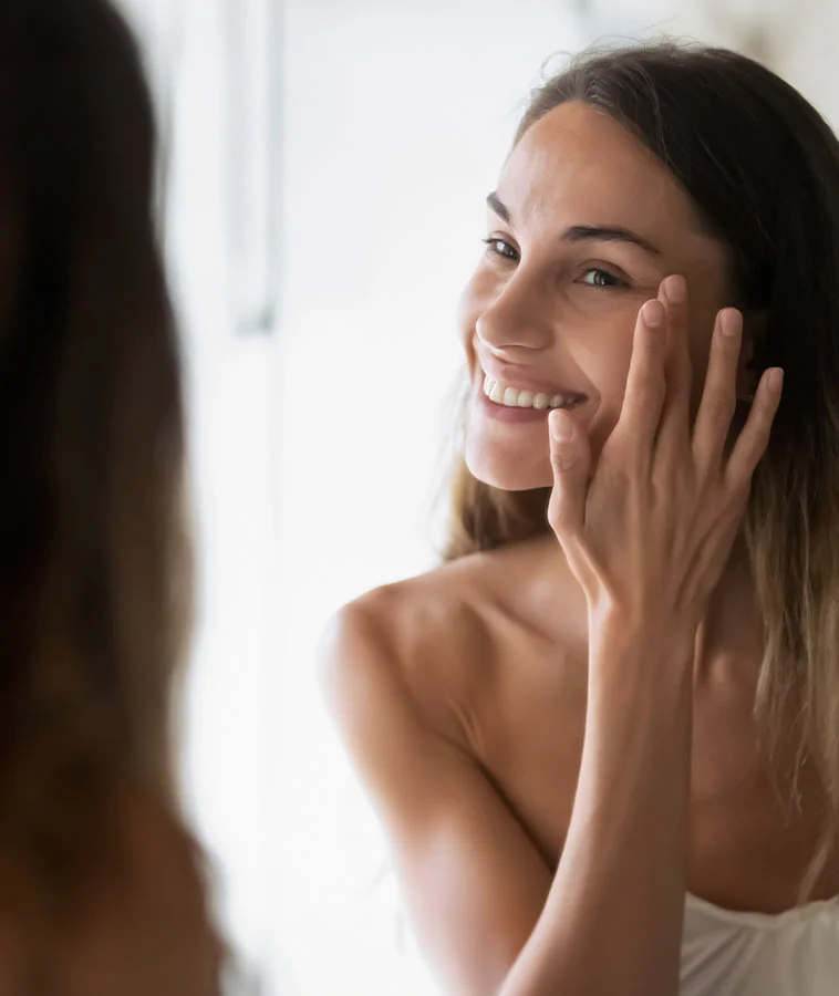 A smiling woman with long hair, touching her face and looking at her reflection in the mirror in a bright, natural light - Eyelid Surgery in New York, NY