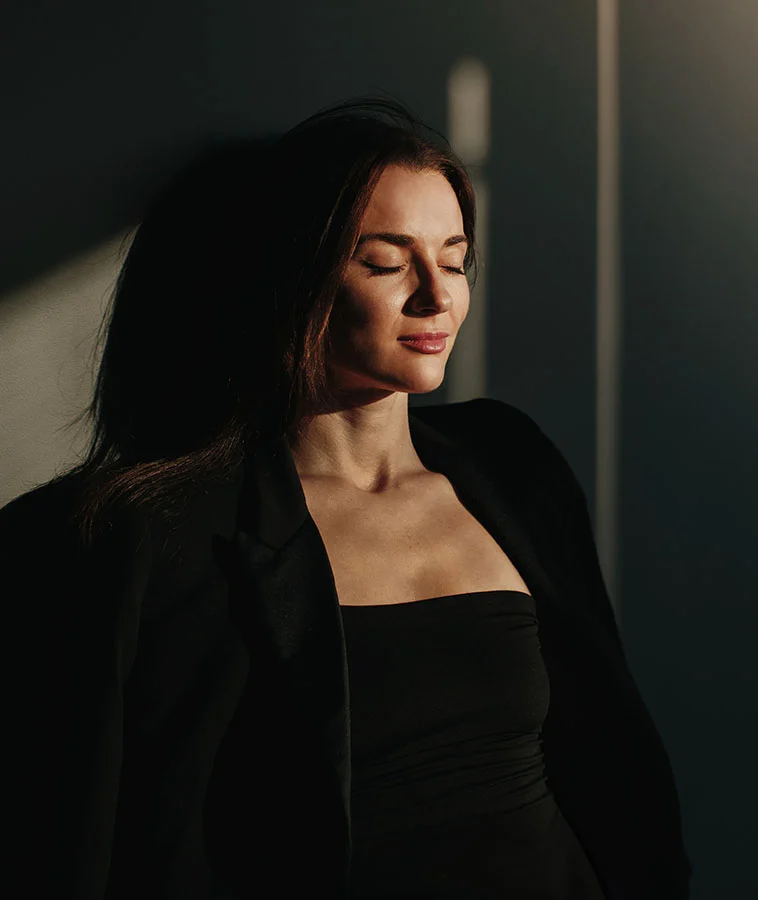 A woman with long dark hair, dressed in a black strapless top and black blazer, standing in dramatic lighting with her eyes closed, basking in sunlight - Fat Grafting Procedure in New York, NY