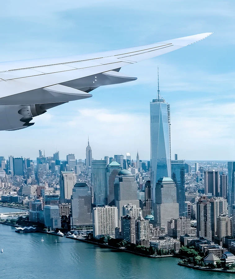 A view from an airplane window, showcasing the aircraft's wing and a panoramic view of a city skyline with tall skyscrapers and a river below - Out of Town Patient Resources in New York, NY